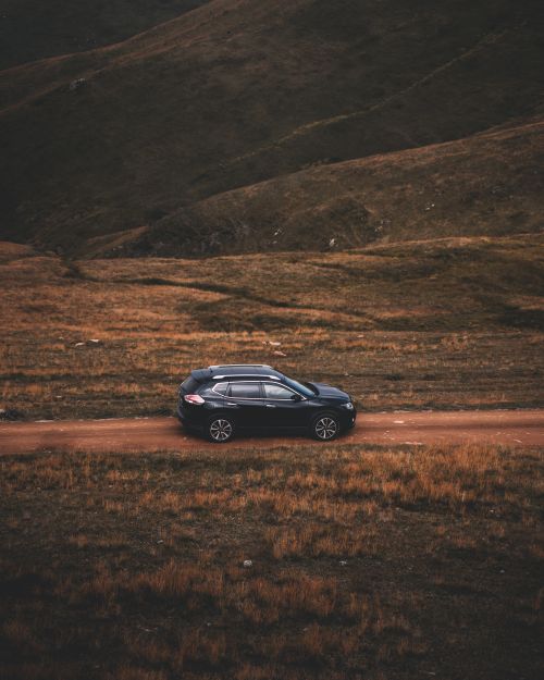 Free A solitary SUV traverses a dirt path through an expansive brown grassy terrain in a mountainous region. Stock Photo