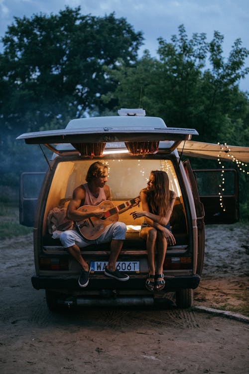 Free Man Playing Guitar Beside a Woman in a Camper Van Stock Photo