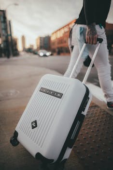 A person with tattoos walks on a city street pulling a white suitcase.