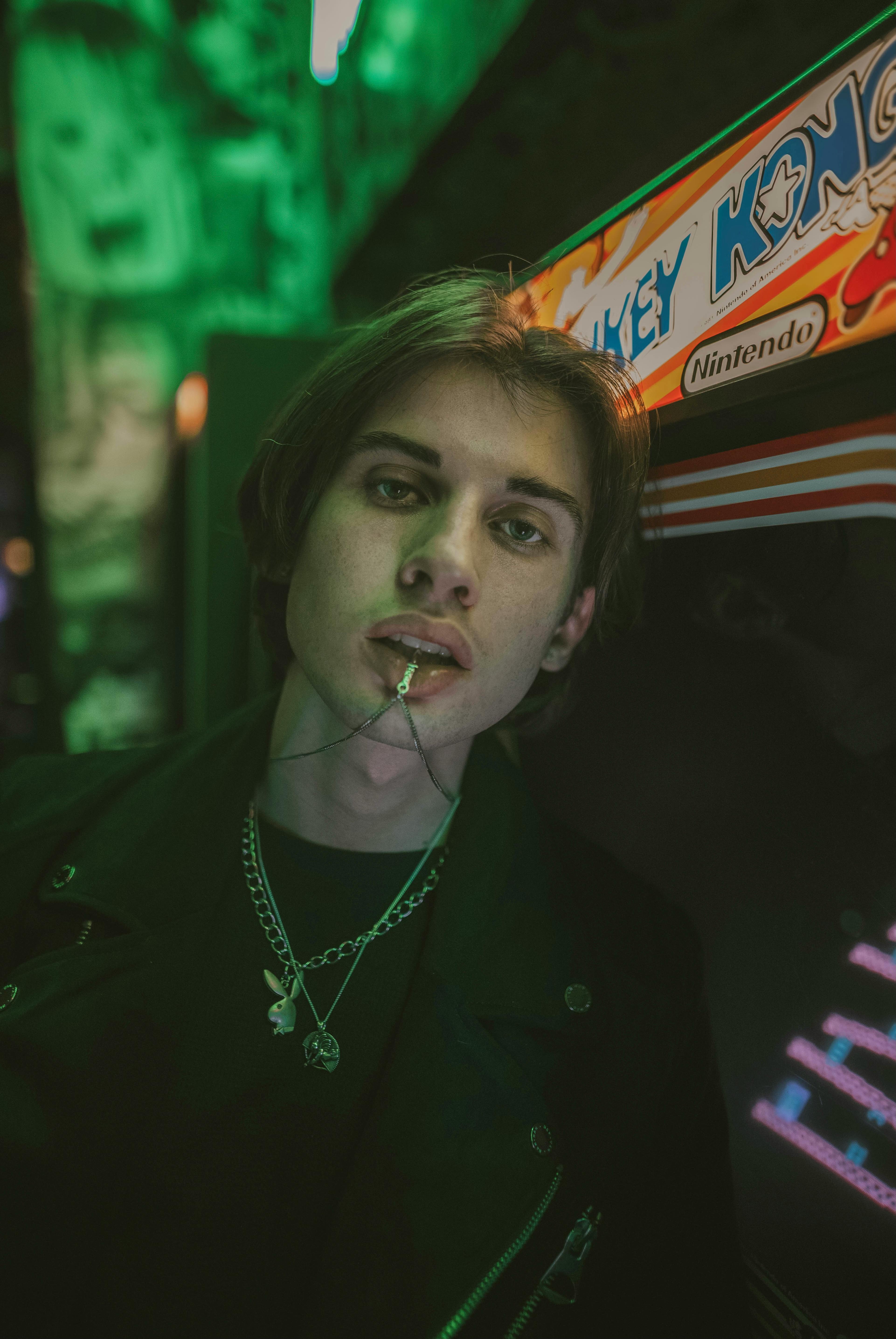 Stylish young man poses in front of a vintage arcade machine with an edgy look and vivid lighting.