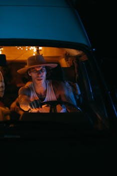 Man driving at night in a van, illuminated by warm string lights, wearing a sunhat.