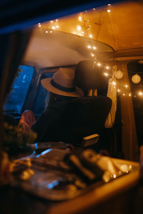 Free A person wearing a hat relaxes inside a warmly lit vintage van adorned with string lights. Stock Photo