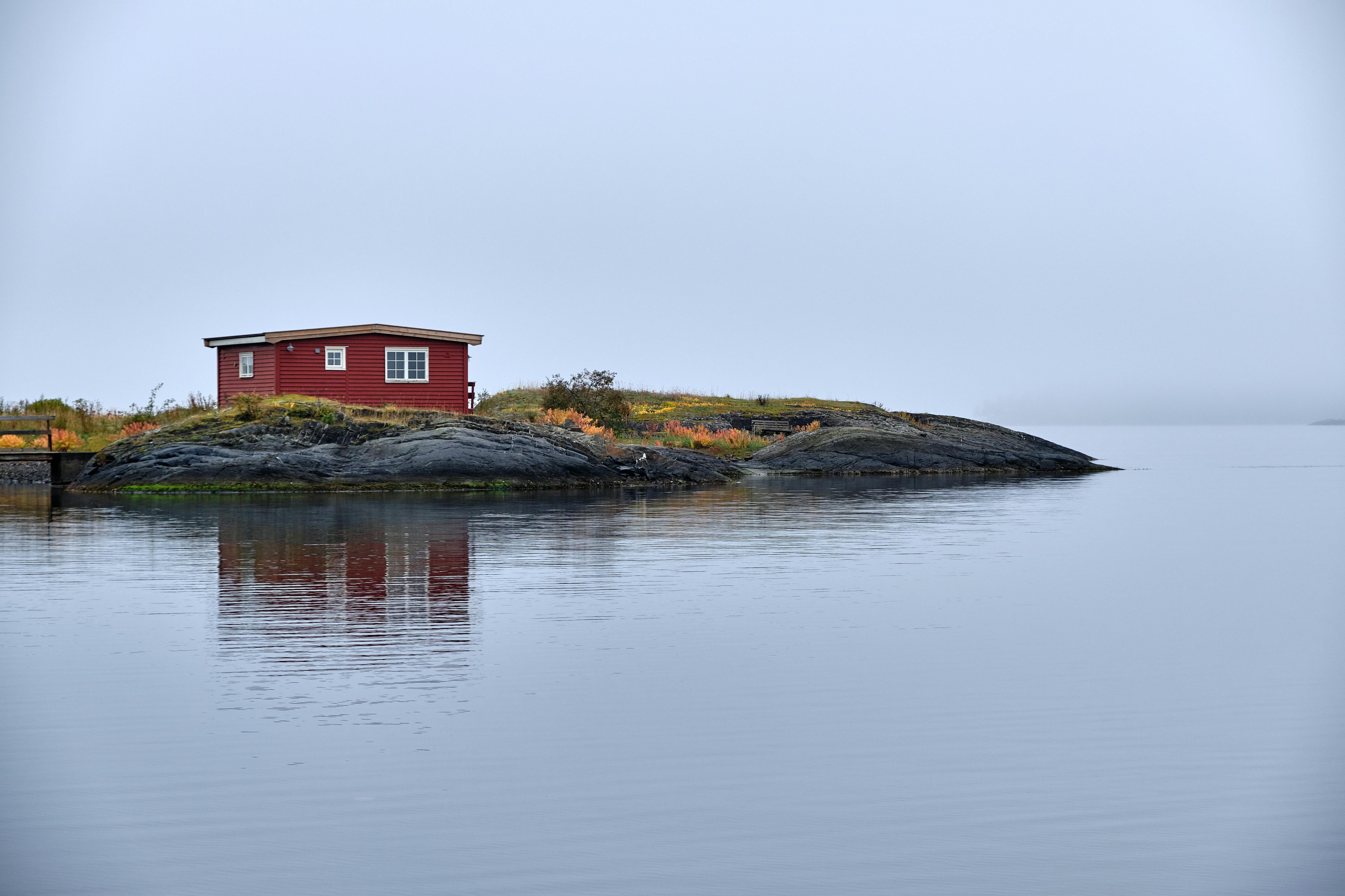 A peaceful red cabin situated on a rocky coastline in Norway, reflecting in calm waters.