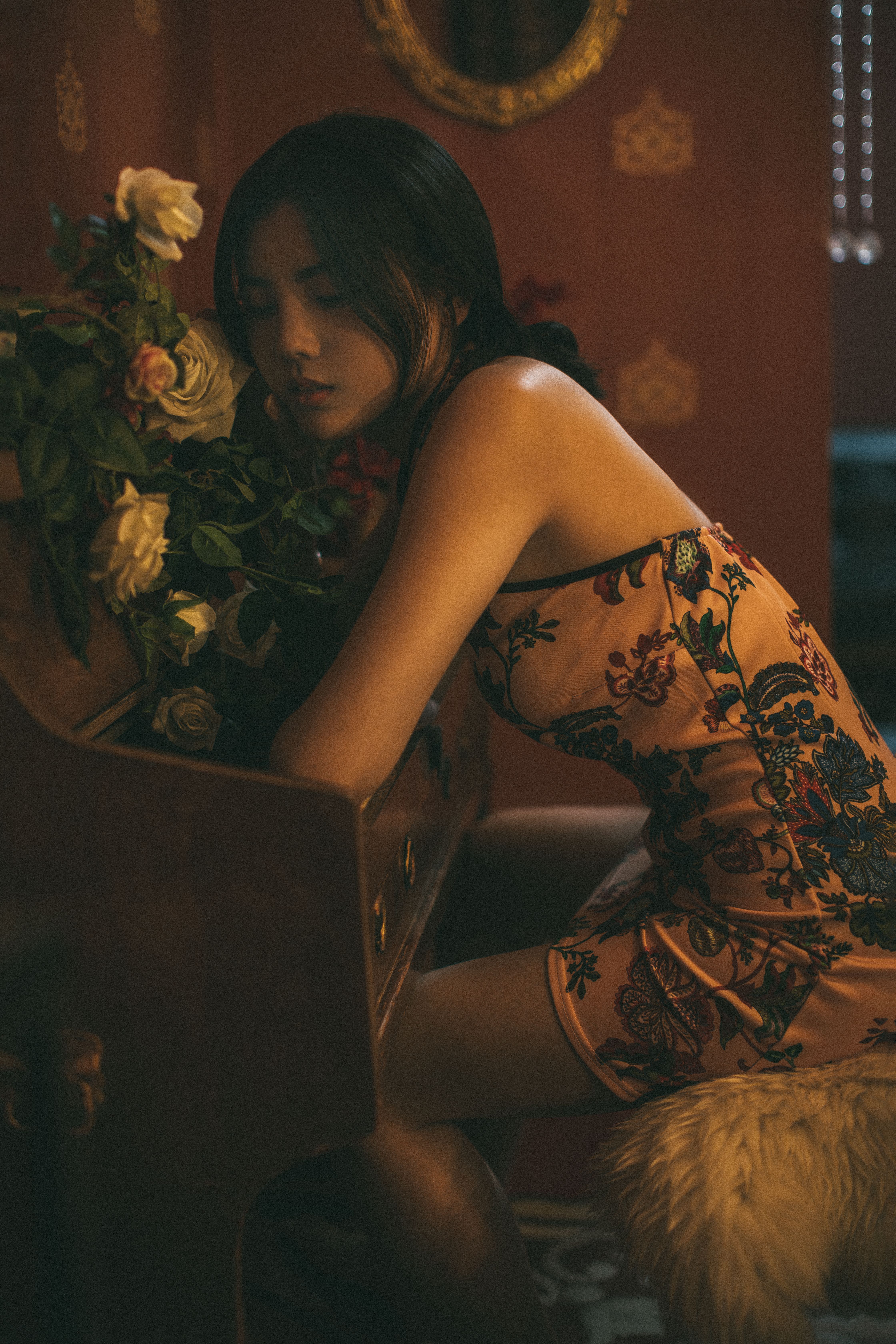 A beautiful woman in embroidered lingerie, sitting in a sultry pose beside a floral arrangement indoors.