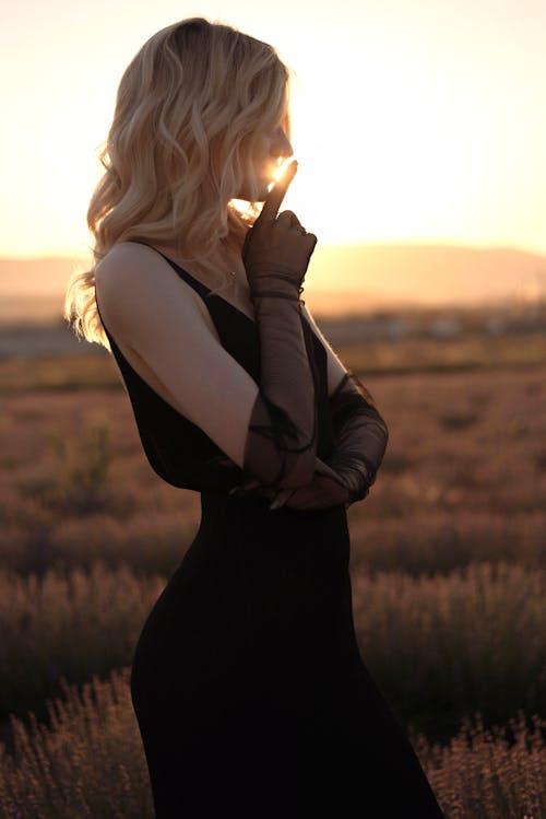 A Woman in Black Dress Standing on Grass Field during Sunset