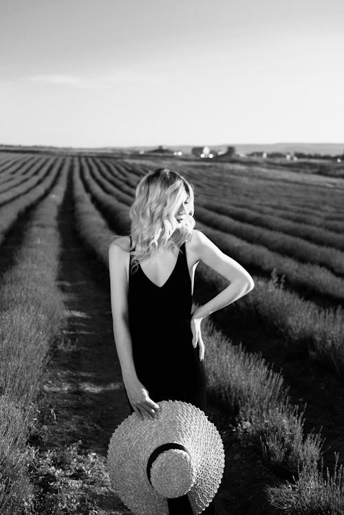 Grayscale Photo of a Woman in a Black Dress Holding a Straw Hat  in a Field