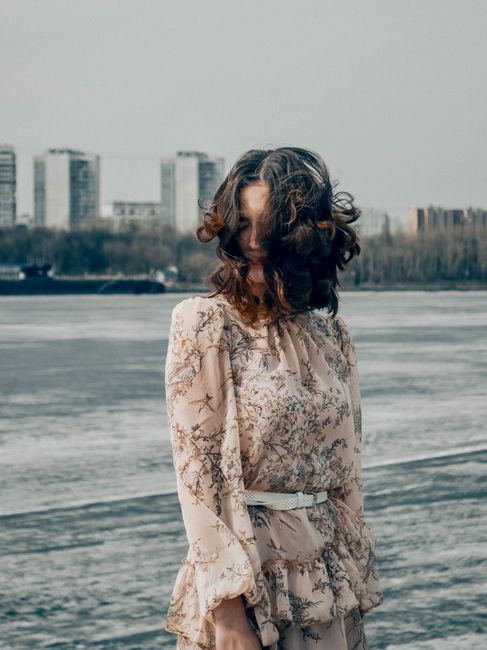A woman in a floral dress by the Moskva River on a windy day, with cityscape in the background.