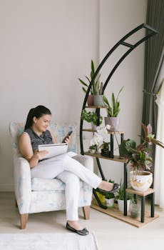 Adult woman sitting in a cozy chair using a smartphone, surrounded by houseplants. Modern lifestyle setting.