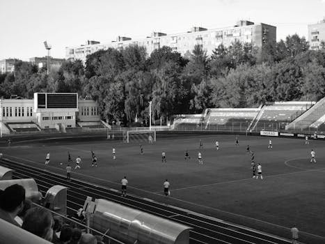 A black and white image of a soccer match taking place in an outdoor stadium with surrounding buildings.