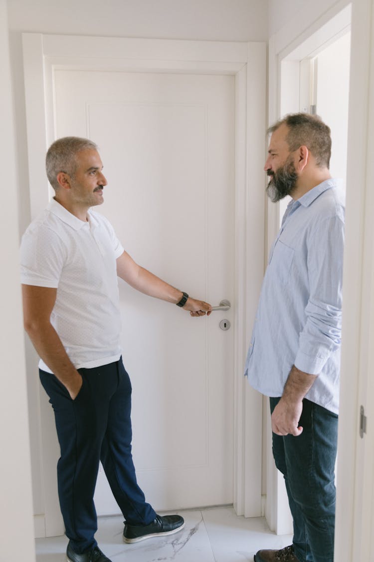 Man In White Polo T-Shirt Holding Door Handle While Talking To Bearded Man In Blue Shirt