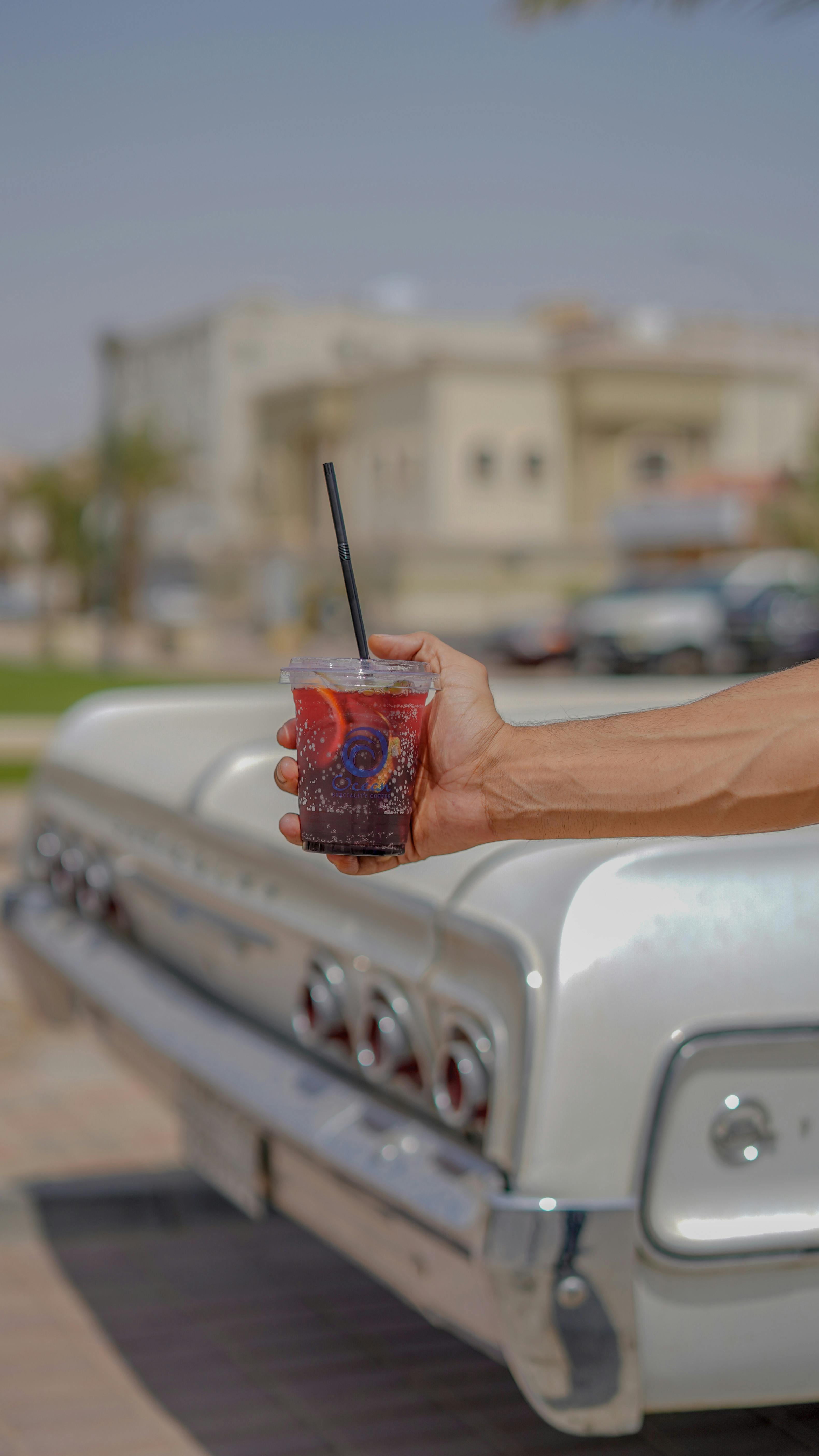 A close-up of a hand holding a cold drink with a straw beside a vintage car.