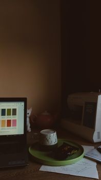 A cozy workspace featuring a laptop, coffee cup, sewing machine, and colorful palette.