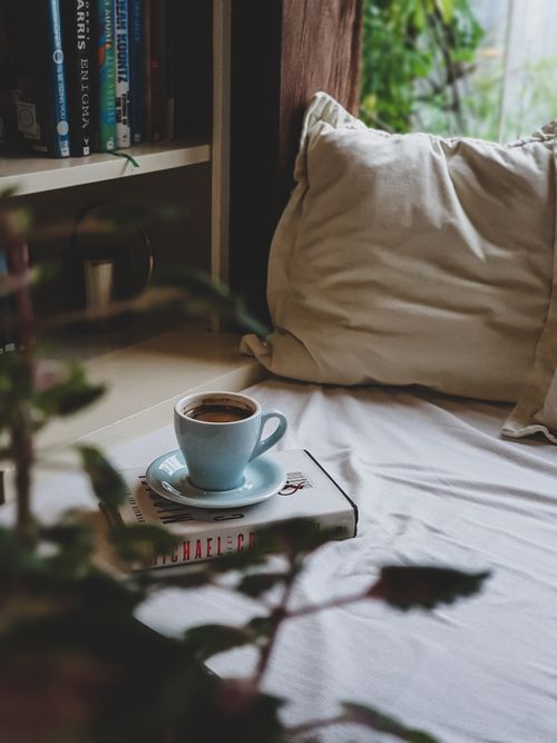 Free A comforting scene featuring a warm cup of coffee resting on a book, set on a cozy bed by a bookshelf. Stock Photo