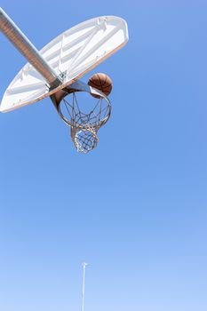 A basketball hoop viewed from below with a ball nearing the net against a clear blue sky.