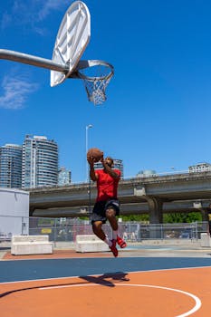 Basketball player performs a layup on outdoor court against city skyline.