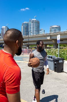 Two men enjoy an outdoor basketball game under a clear blue sky in an urban setting.