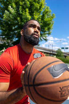 Bearded man in red shirt holding a basketball outdoors with trees and blue sky.