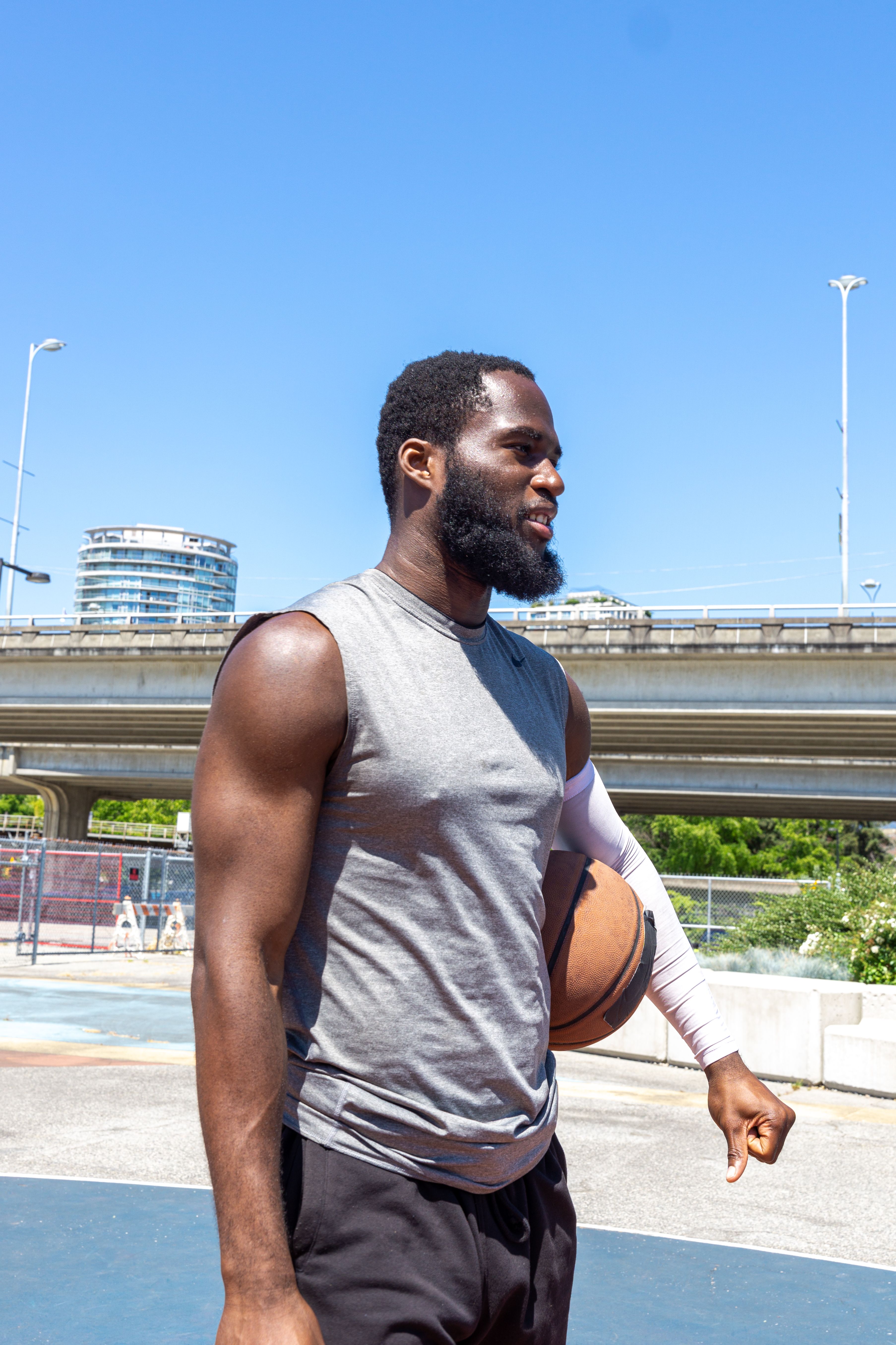 A young man stands confidently with a basketball on an outdoor court under a clear blue sky.