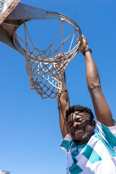 Smiling young man hanging from a basketball hoop under a clear blue sky.