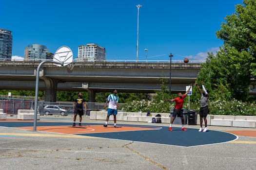 Four men playing basketball on an outdoor court under a city overpass on a sunny day.