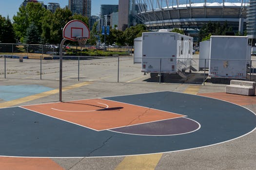Outdoor basketball court in urban setting, sunny day, city skyline and stadium visible.