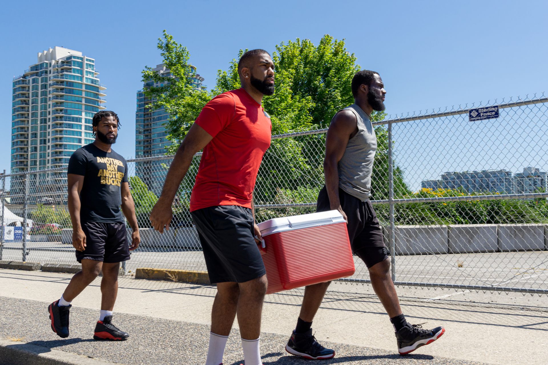 Three men casually walking outdoors holding a cooler, enjoying a sunny day.