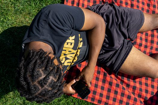 Adult male with braids using smartphone while lying on checkered picnic blanket outdoors.