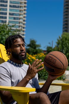 A man sitting outdoors in a chair holding a basketball on a sunny day.