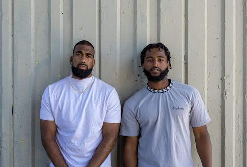 Two Men Standing beside the Gray Wall while Looking at Camera