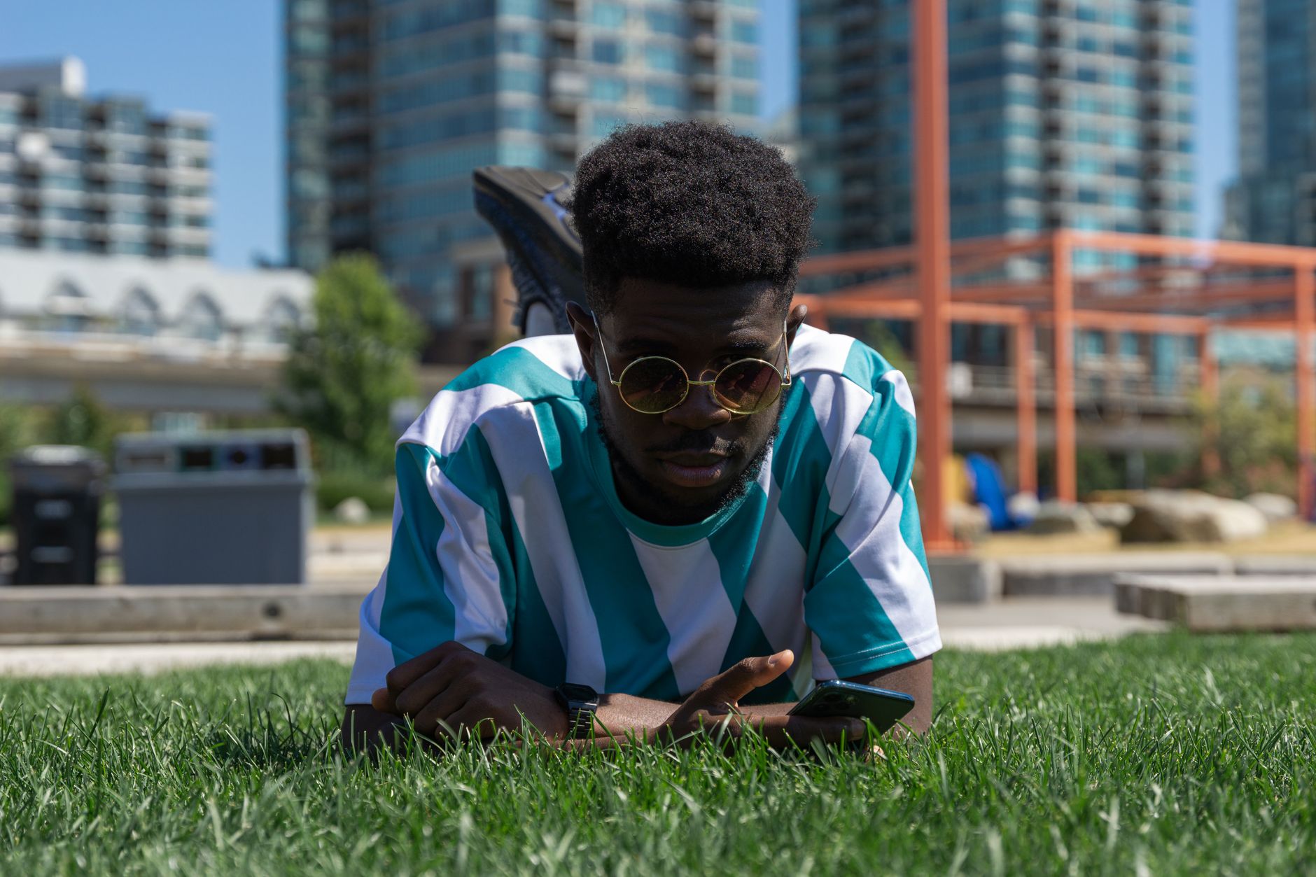 An adult male enjoys modern technology in an urban park setting, lying on grass.