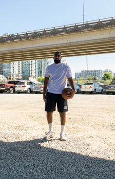 Man holding a basketball under an urban bridge on a sunny day wearing activewear.