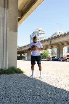 Athlete wearing sportswear holds a basketball under a city bridge, bathed in sunlight.