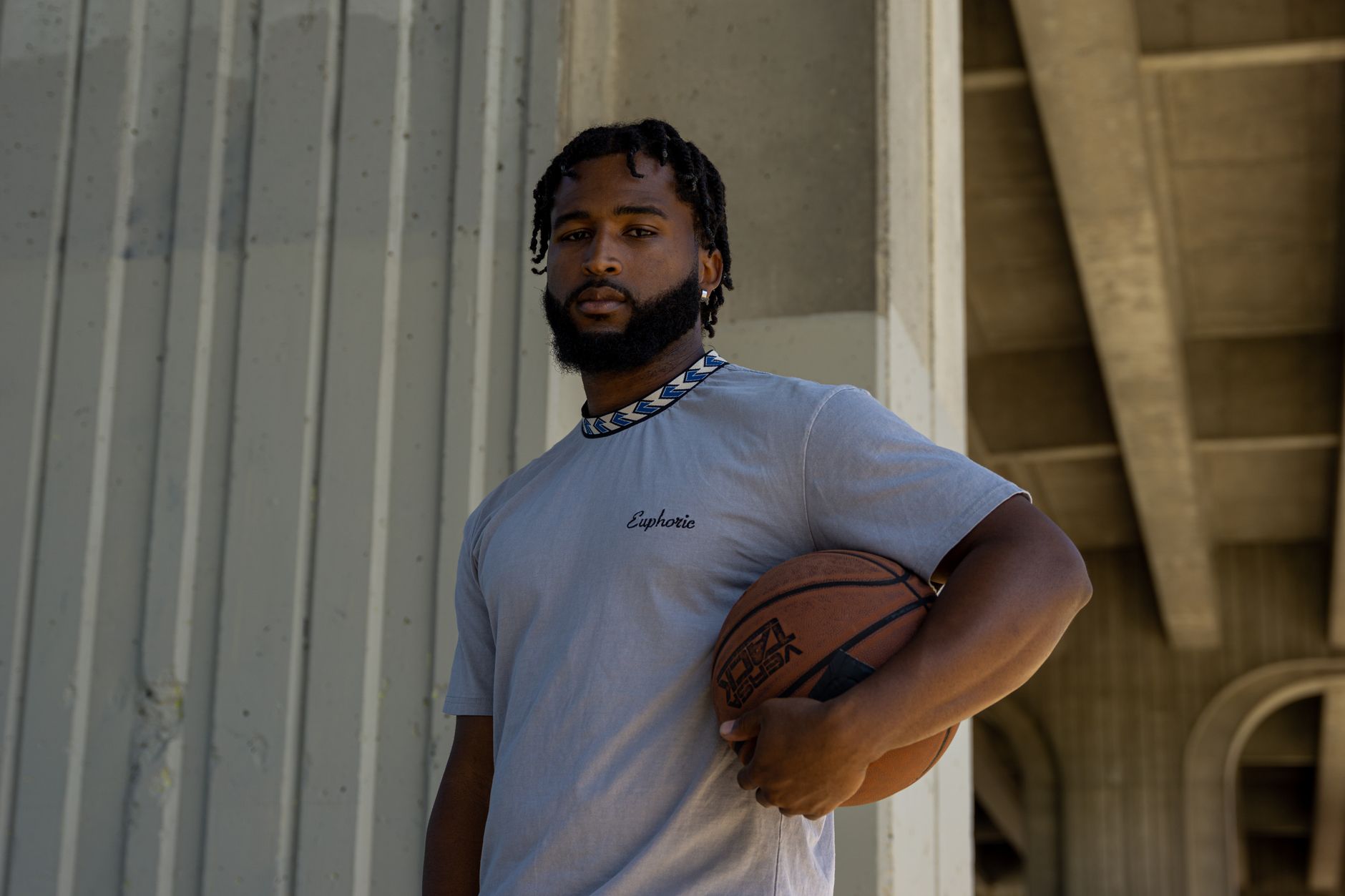 Portrait of a man with dreadlocks holding a basketball, standing confidently under a bridge.