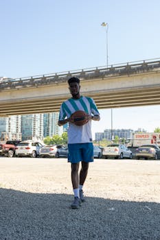 A man in sportswear holding a basketball under a bridge with urban background on a sunny day.