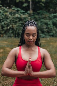A woman with braided hair meditating outdoors in serene nature setting, wearing red activewear.