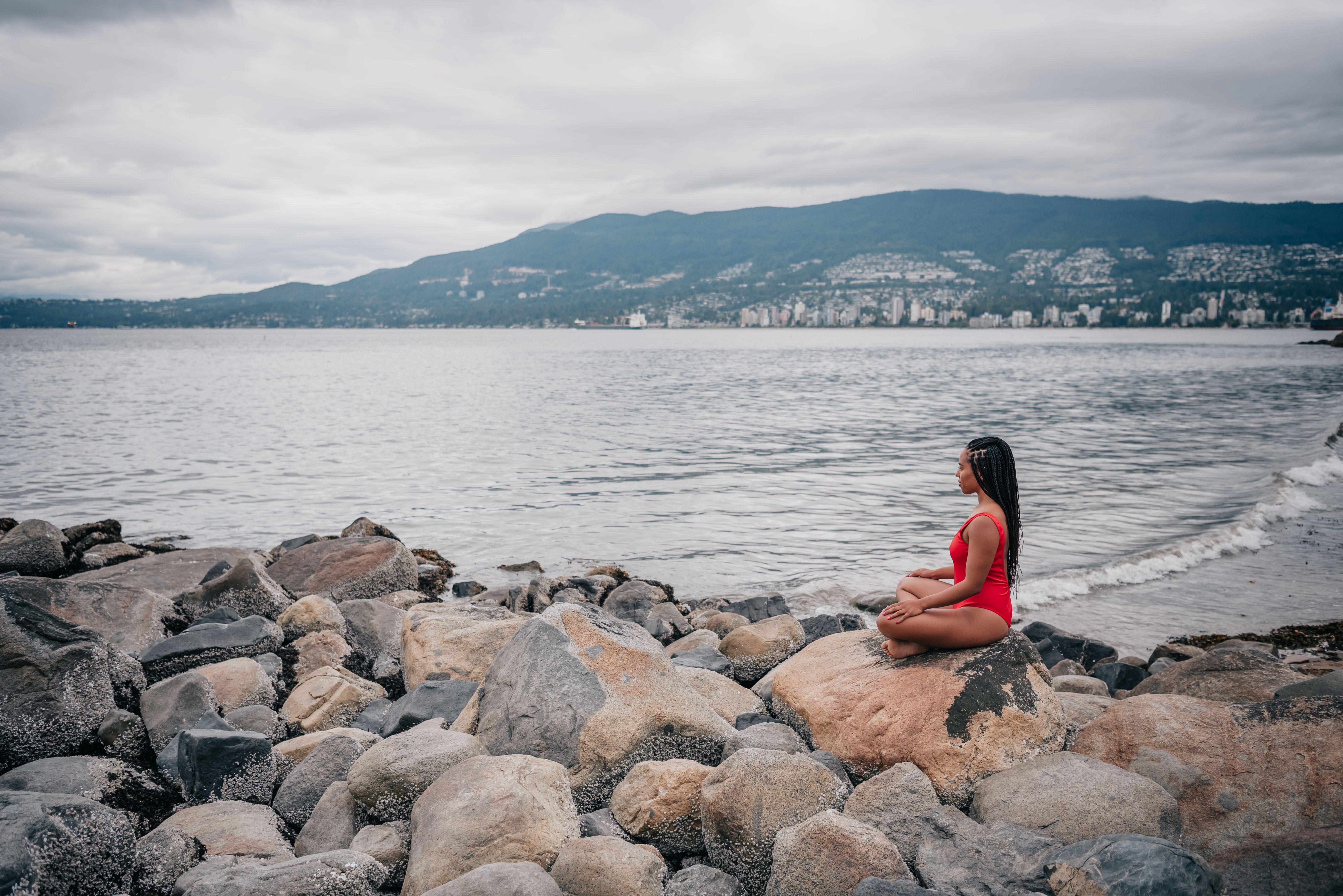 A woman in a red swimsuit meditates on a rocky shore by the ocean under a cloudy sky.