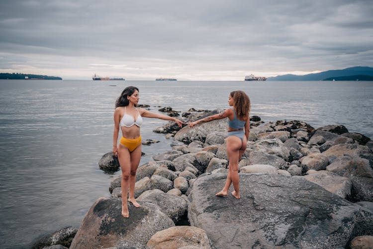 Women In Bikinis Touching Fingers While Standing On A Rocky Shore