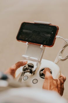 Hands holding a drone controller connected to a smartphone, showing real-time video feed.