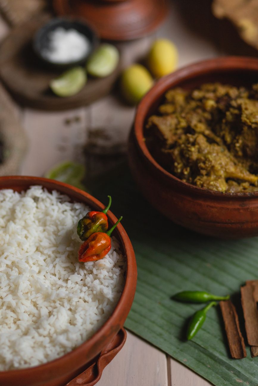 Bowl of dark reddish-brown goat curry with tender meat and potato chunks, served alongside white rice with visible scotch bonnet peppers