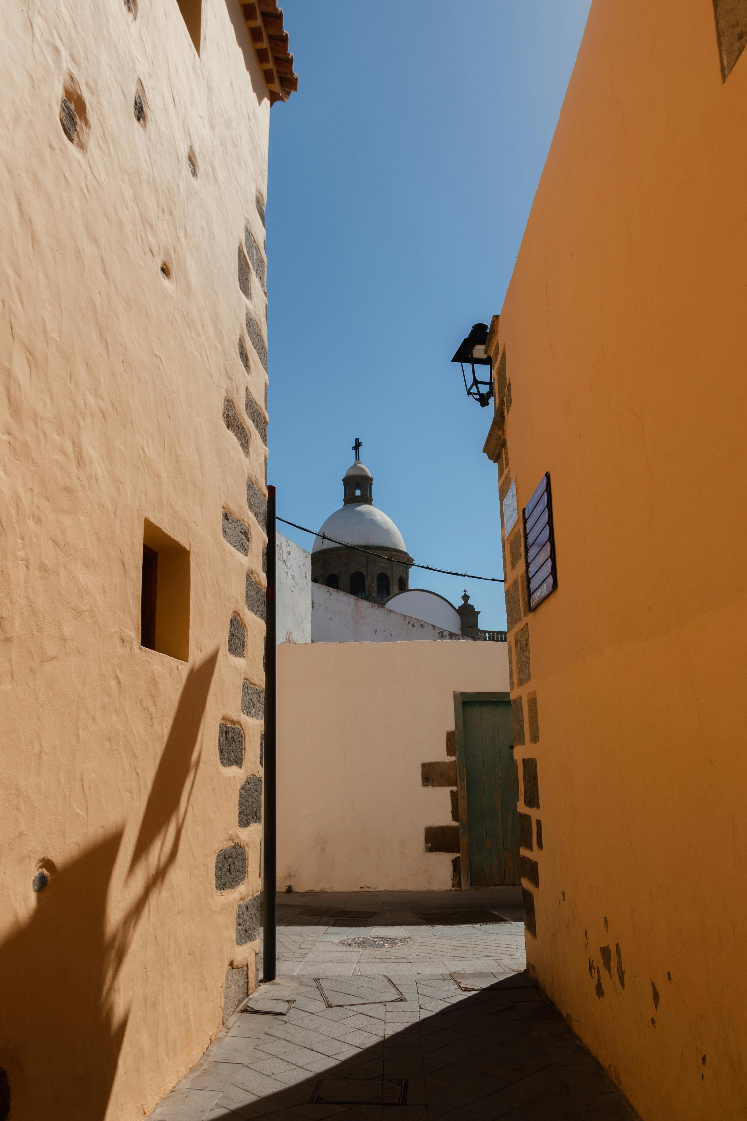 Narrow alleyway with colorful walls leading to a historic church dome in Spain, under a clear blue sky.