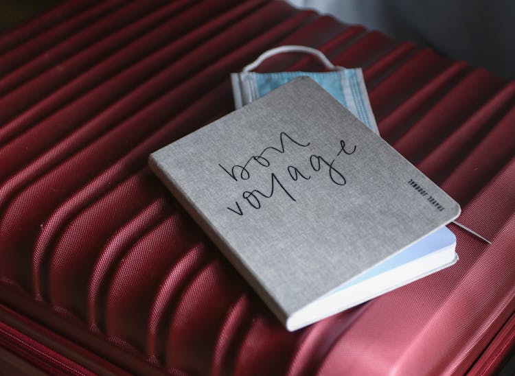 A Gray Notebook And Face Mask On  Red Suitcase 