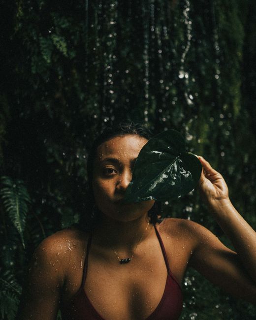 Woman with a leaf in front of face next to a waterfall, serene natural setting.