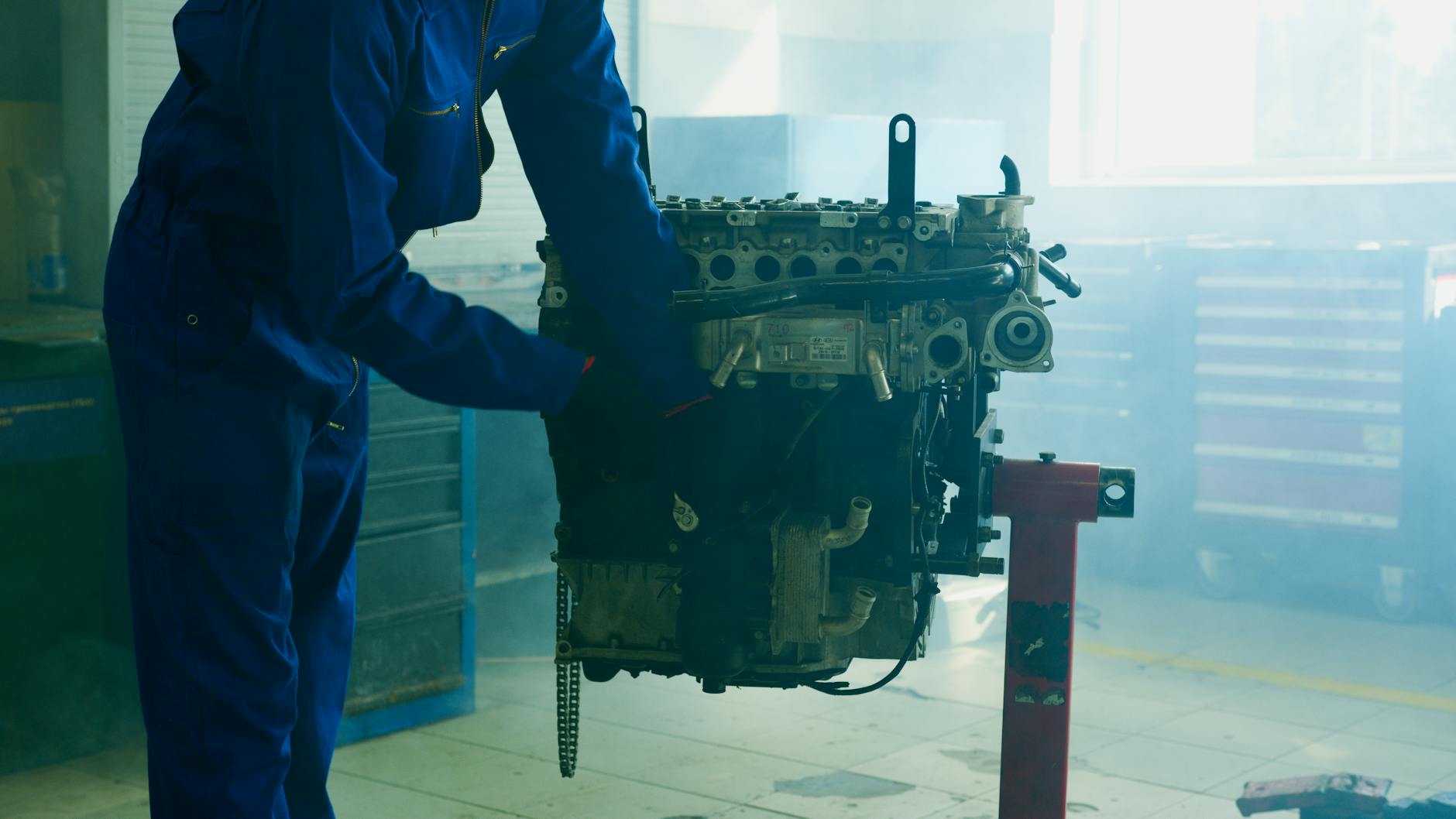 Mechanic in blue overalls inspecting an engine in an indoor garage.