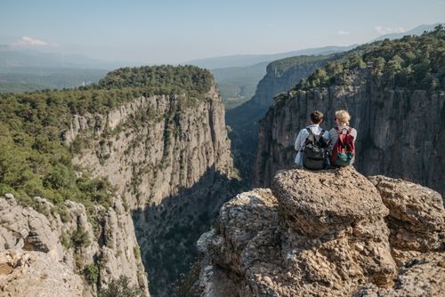 Free Two people sitting on a cliff edge, capturing a breathtaking view of a rocky canyon surrounded by lush forest. Stock Photo