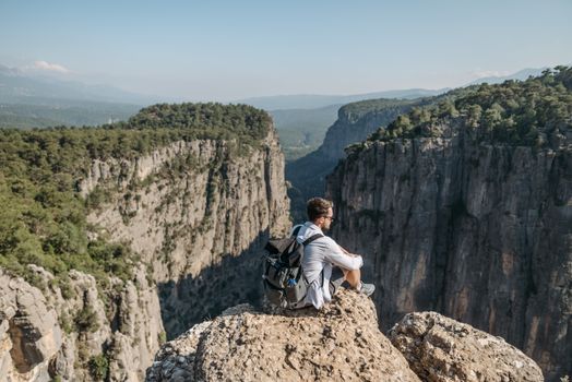 A man sitting on a rocky edge overlooking a dramatic mountain canyon under a clear sky.