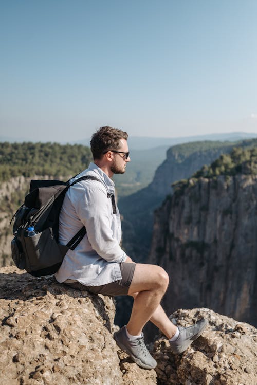 Man in Denim Shirt and Backpack Sitting on the Rock on Top of the Cliff