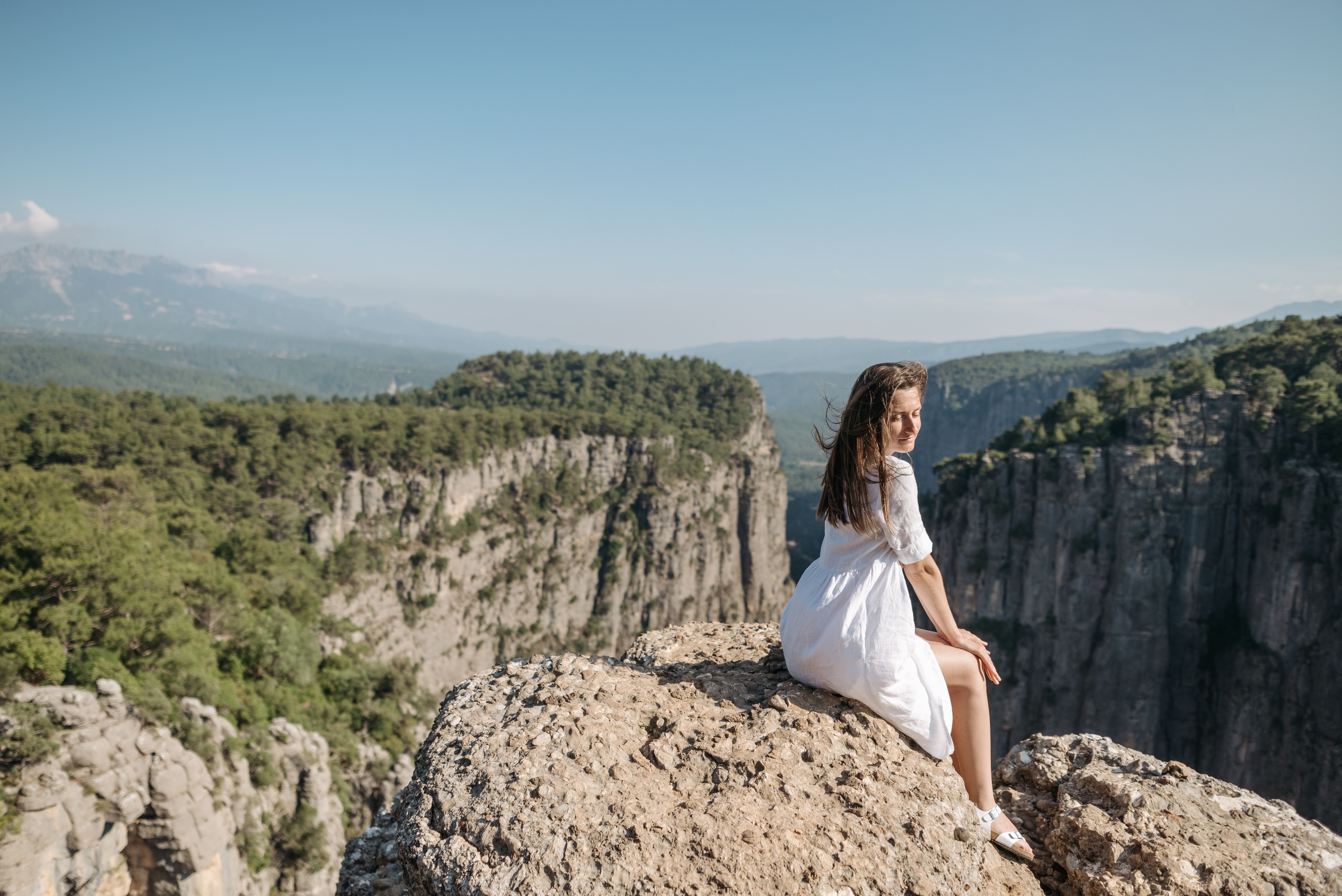 A woman in a white dress sitting on a cliff, overlooking a vast forest and distant mountains on a sunny day.