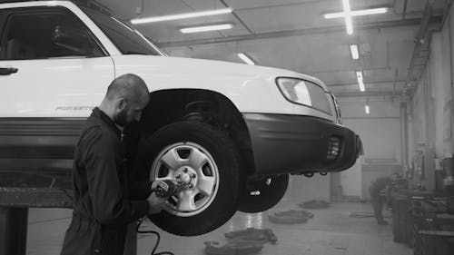 Grayscale Photo of a Mechanic Using an Impact Wrench on a Car in an Auto Repair Shop