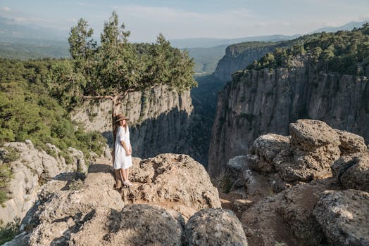 A woman in a white dress stands on rocky terrain by a canyon, wearing a hat, under bright sunlight.