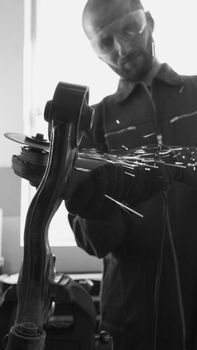 Black and white image of an auto mechanic using an angle grinder on a car part, indoors with sparks flying.